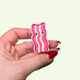 Hand holding a pink bacon-shaped ring with a smiley face on a light beige background