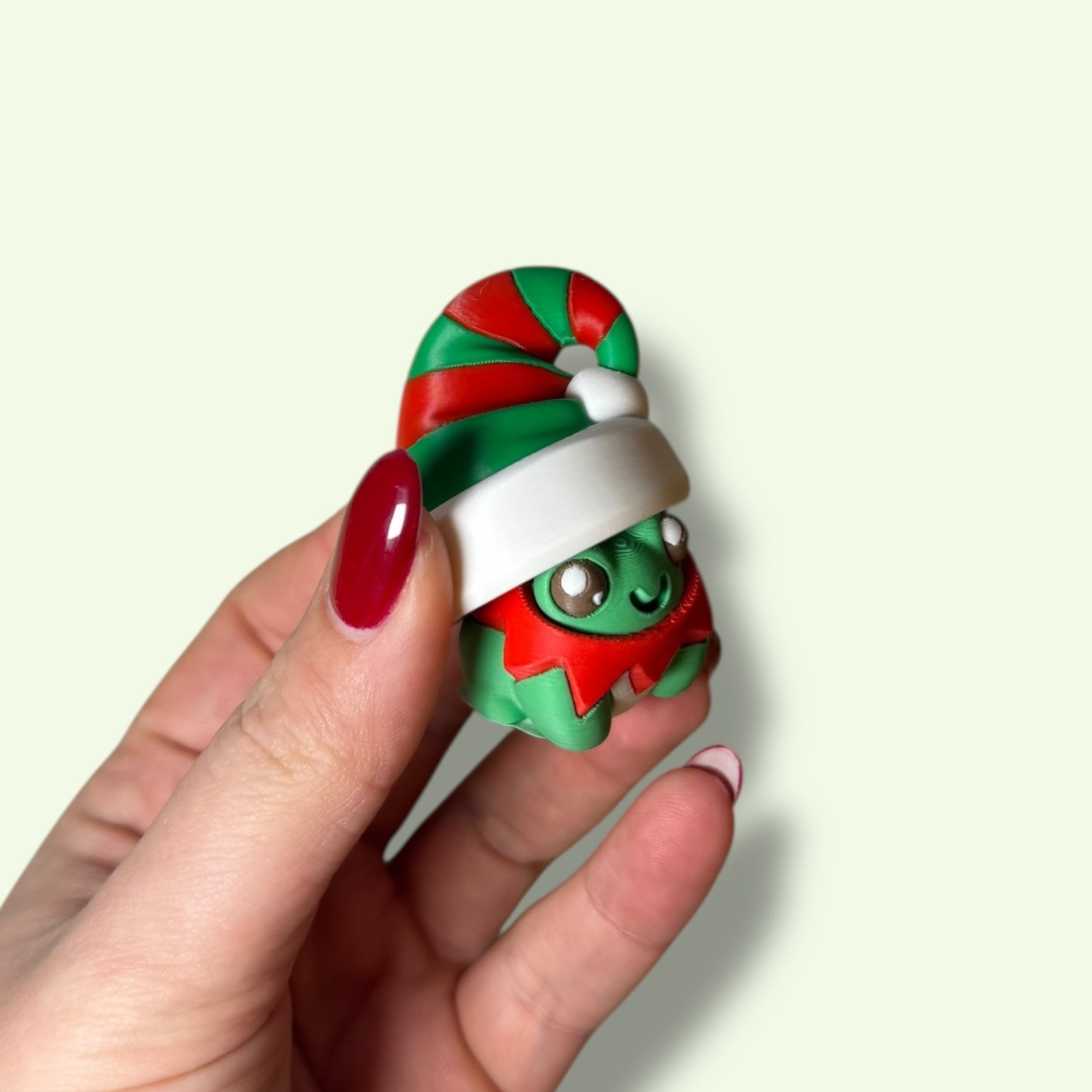 Hand holding a small green creature with red and white accents against a light background